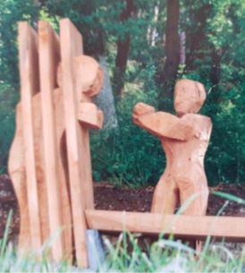 Zwei Holzfiguren: eine hinter Gittern, eine zweite außerhalb streckt die Arme nach der Figur hinter Gittern aus. Objektbank am Besinnungsweg Hainsacker.