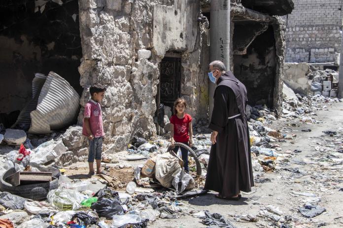 Pater Firas steht mit 2 Kindern in einem Ruinenfeld
