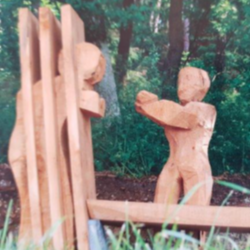 Zwei Holzfiguren: eine hinter Gittern, eine zweite außerhalb streckt die Arme nach der Figur hinter Gittern aus. Objektbank am Besinnungsweg Hainsacker.